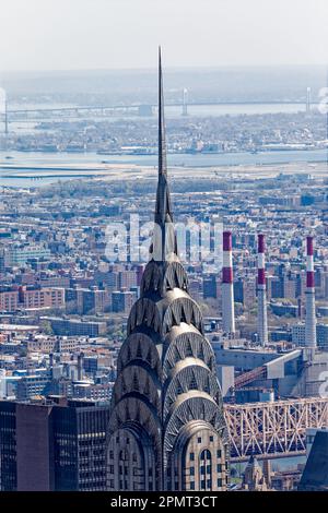 Blick von oben: Der Edelstahlspieß des New York City Icon Chrysler Building leuchtet unter der Mittagssonne; der Stadtteil Queens liegt dahinter. Stockfoto