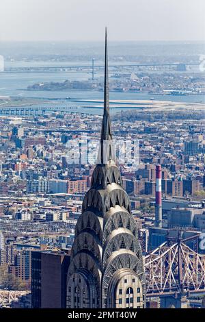 Blick von oben: Der Edelstahlspieß des New York City Icon Chrysler Building leuchtet unter der Mittagssonne; der Stadtteil Queens liegt dahinter. Stockfoto