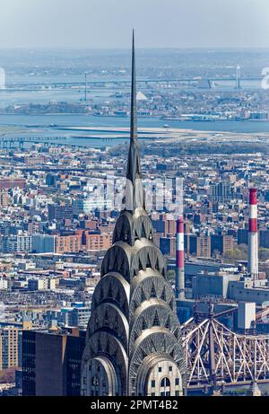 Blick von oben: Der Edelstahlspieß des New York City Icon Chrysler Building leuchtet unter der Mittagssonne; der Stadtteil Queens liegt dahinter. Stockfoto