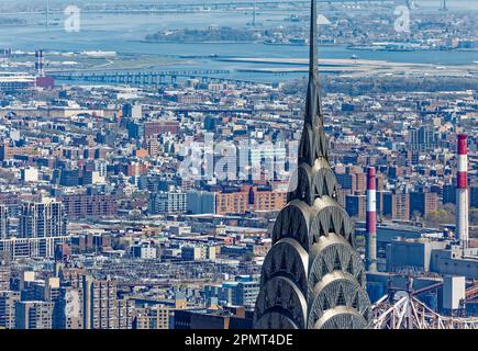 Blick von oben: Der Edelstahlspieß des New York City Icon Chrysler Building leuchtet unter der Mittagssonne; der Stadtteil Queens liegt dahinter. Stockfoto