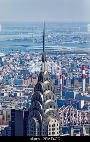 Blick von oben: Der Edelstahlspieß des New York City Icon Chrysler Building leuchtet unter der Mittagssonne; der Stadtteil Queens liegt dahinter. Stockfoto