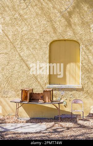 Attraktives geschlossenes Fenster auf einem gelben Stuckgebäude mit langen Schatten. Vor dem Hotel befindet sich ein abfallender Tisch mit einem alten Schreibtisch neben einem Metallstuhl. Stockfoto