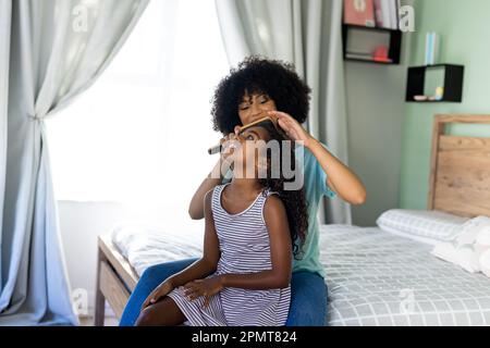Fröhliche, birassische Mutter, die ihrer Tochter die Haare bürstet, während sie im Bett im Schlafzimmer sitzt Stockfoto