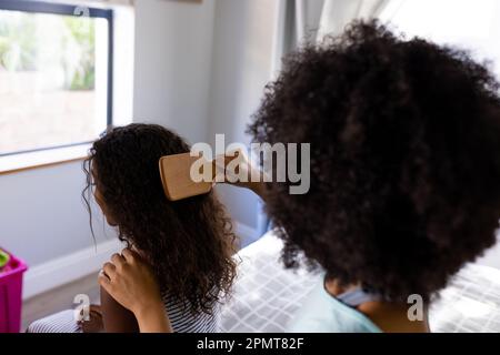 Rückansicht einer birassischen Mutter, die ihrer Tochter die Haare bürstet und im Schlafzimmer auf dem Bett sitzt Stockfoto