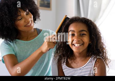 Porträt einer lächelnden, birassischen Tochter mit Mutter, die sich im Schlafzimmer die Haare bürstet Stockfoto