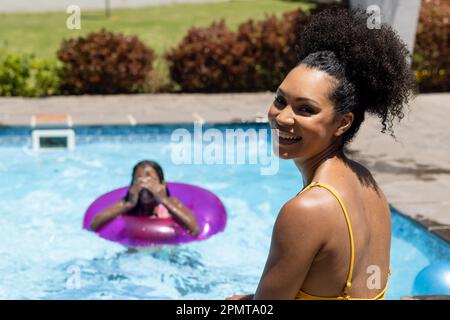 Porträt einer fröhlichen, birassischen Mutter, die lächelt, während die Tochter im sonnigen Außenpool spielt Stockfoto