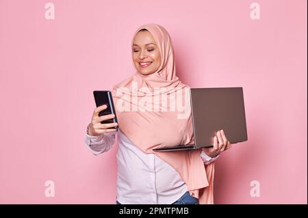 Arabische muslimische Frau, arbeitende schwangere Geschäftsfrau in rosa Hijab, mit Handy und Laptop. Schwangerschaft und Karriere Stockfoto