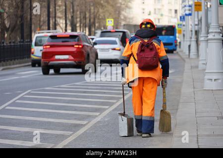 Eine Arbeiterin in orangefarbener Uniform mit Besen fegt die Straße auf dem Hintergrund von Autos. Straßenreinigung in der Frühjahrsstadt Stockfoto