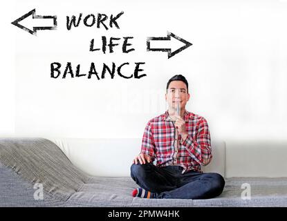 Die Worte Work Life Balance, geschrieben an einer Wand, mit Pfeilen in entgegengesetzte Richtungen, und ein Mann, der auf einer Couch sitzt und sie ansieht Stockfoto