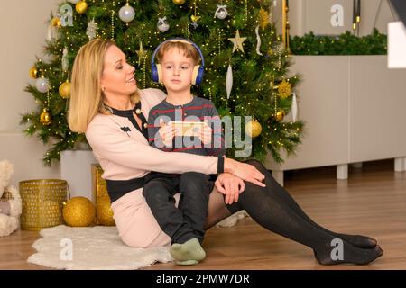 Der fünfjährige Sohn sitzt auf dem Schoß seiner Mutter, spielt mit seinem Handy und die Mutter sieht ihn vor dem dekorierten Weihnachtsbaum an Stockfoto