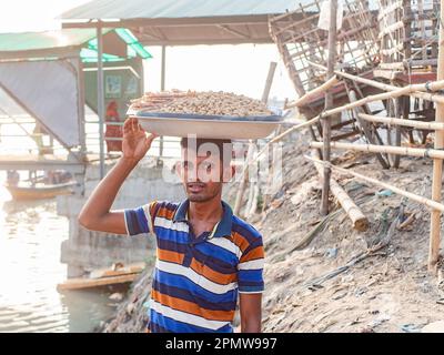 Ein Mann, der ein Tablett mit Erdnüssen zu lokalen Fähren am Wise Ghat Boat Station am Buriganga River in Dhaka, der Hauptstadt von Bangladesch, transportiert. Stockfoto