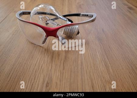 Gebrauchte Glühlampe und Schutzbrille aus Kunststoff auf Holzboden. Austausch der verschmutzten Glühlampe während der Renovierung. Stockfoto