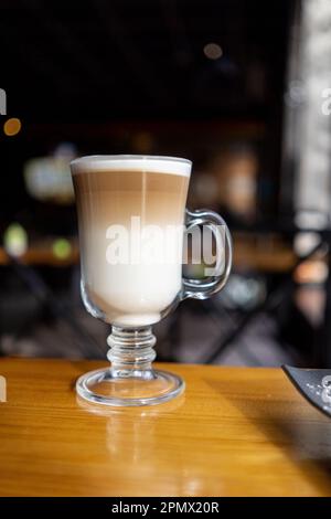 Cafe Latte Macchiato geschichteten Kaffee in einem See-through-Glas irish Coffee Glas. Die Tasse steht auf einem Holztisch mit Kaffeebohnen auf dem Tisch neben dem Stockfoto