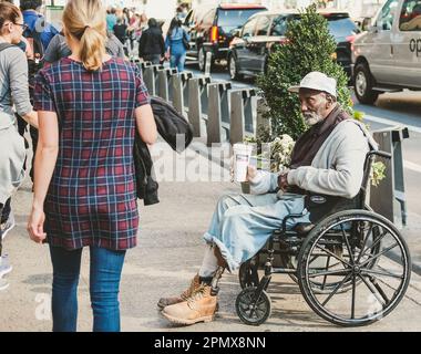 Ein behinderter Mann, der nach Wohltätigkeit fragt, während er im Rollstuhl sitzt, während die Leute vorbeigehen. Der Mann ist mit einem künstlichen linken Unterschenkel zu sehen. Stockfoto