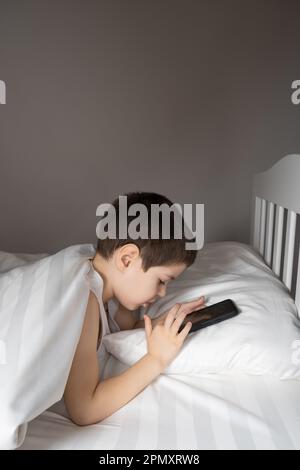 Ein 5-jähriger Junge spielt Spiele auf seinem Handy, während er im Bett liegt. Geräte im Bett vor dem Schlafengehen bei einem Kind Stockfoto