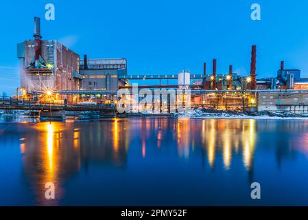 In der Abenddämmerung wirft die beleuchtete Fabrik eine Reflexion auf den Fluss. Lampen beleuchten dieses markante Industriegebäude. Stockfoto