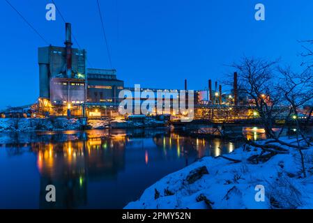 In der Abenddämmerung wirft die beleuchtete Fabrik eine Reflexion auf den Fluss. Lampen beleuchten dieses markante Industriegebäude. Stockfoto