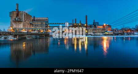 In der Abenddämmerung wirft die beleuchtete Fabrik eine Reflexion auf den Fluss. Lampen beleuchten dieses markante Industriegebäude. Stockfoto