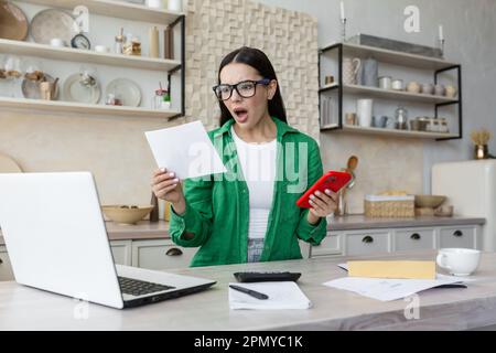 Schockierte junge Frau, die einen Brief las, während sie zu Hause in der Küche saß. Er hält das Telefon in den Händen, sieht sich die Dokumente besorgt an. Stockfoto