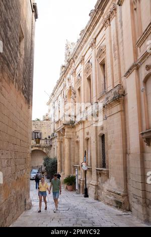 Mdina, Malta - 13. November 2022: Kalksteingebäude in den gepflasterten Straßen der ruhigen Stadt und ein Touristenpaar, das zu Fuß unterwegs ist Stockfoto