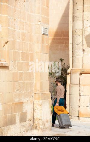 Mdina, Malta - 13. November 2022: Mann, der auf der Fußgängerzone durch Kalksteine läuft, während er seinen Koffer von hinten zieht Stockfoto