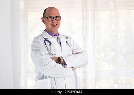 Porträt eines männlichen Arztes mit weißem Mantel, lila Hemd, Krawatte, Uhr und Brille. Ein Stethoskop hängt an seinem Hals. Ein Fenster ist auf d Stockfoto
