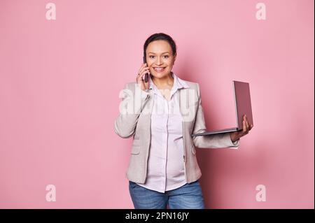 Stilvolle, schwangere Geschäftsfrau, die mit dem Handy spricht, mit Geschäftspartnern verhandelt, mit dem Laptop auf Pink posiert Stockfoto