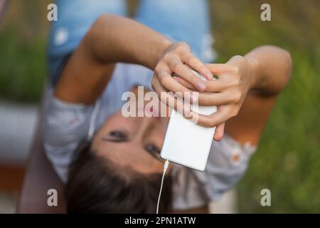 Eine junge Frau mit braunem Haar macht Selfies oder führt mit ihrem weißen Smartphone Videoanrufe. Sie streckt ihre Arme aus, um das Gestell beim Tragen zu verbessern Stockfoto