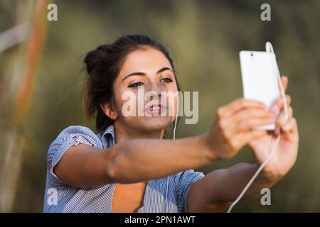 Eine junge Frau mit braunem Haar macht Selfies oder führt mit ihrem weißen Smartphone Videoanrufe. Sie streckt ihre Arme aus, um das Gestell beim Tragen zu verbessern Stockfoto