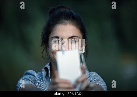 Eine junge Frau mit braunem Haar macht Selfies oder führt mit ihrem weißen Smartphone Videoanrufe. Sie streckt ihre Arme aus, um das Gestell beim Tragen zu verbessern Stockfoto