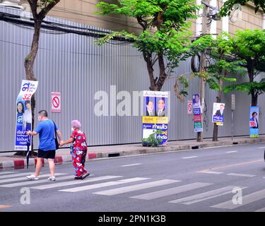 Ein Mann hilft einer älteren Frau über die Surawong Road. Wahlkandidaten verwenden Plakate an Straßenmöbeln, um sich bei potenziellen Wählern im Silom District, im Zentrum von Bangkok, Thailand und Südostasien zu bewerben. Nach der Auflösung des 25. Repräsentantenhauses am 20. März 2023 werden in Thailand am 14. Mai 2023 allgemeine Wahlen stattfinden. Stockfoto