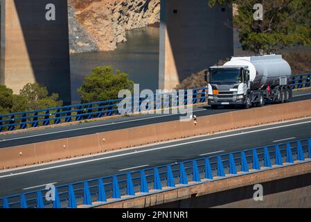 Tankwagen, der mit den Säulen einer Brücke hinter der Autobahn fährt. Seitenansicht. Stockfoto