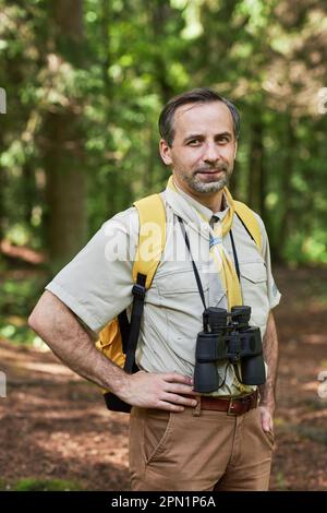 Vertikales Porträt eines erwachsenen Mannes als Scout-Führer, der im Wald draußen auf die Kamera schaut und Wanderausrüstung trägt Stockfoto