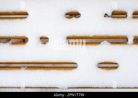 Blick von oben auf die schneebedeckte gelbe Holzbank aus nächster Nähe im Stadtpark am bedeckten Wintertag Stockfoto