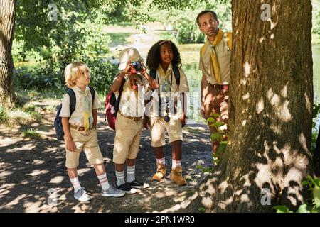 Vielfältige Gruppe kleiner Pfadfinder im Wald, die die Natur erkunden und in Ferngläsern schauen Stockfoto