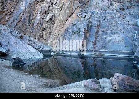 Reflexionen im Pool unter einem trockenen Wasserfall, umgeben von Felsen mit schrägen Felsen auf der linken Seite. Stockfoto