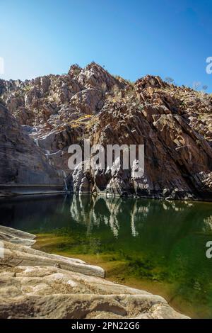 Reflexionen im Pool unter einem trockenen Wasserfall, umgeben von Felsen mit grünem Wasserunkraut, das wächst. Stockfoto