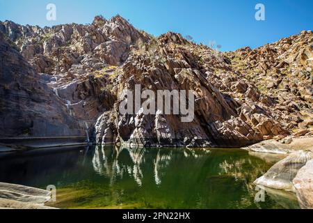 Reflexionen im Pool unter einem trockenen Wasserfall, umgeben von Felsen mit grünem Wasserunkraut, das wächst. Stockfoto