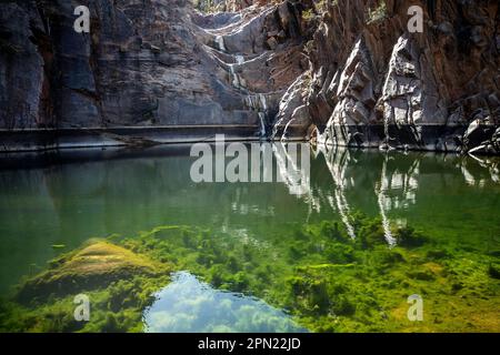 Reflexionen im Pool unter einem trockenen Wasserfall, umgeben von Felsen mit grünem Wasserunkraut, das wächst. Stockfoto