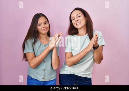 Junge Mutter und Tochter stehen über rosa Hintergrund und klatschen und applaudieren fröhlich und fröhlich, lächeln stolze Hände zusammen Stockfoto