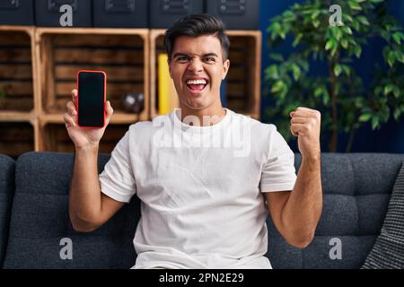 Ein hispanischer Mann mit einem Smartphone zeigt einen leeren Bildschirm, der stolz schreit und den Sieg und Erfolg mit erhobenen Armen feiert Stockfoto