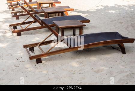 Leere verstellbare Holzliegen ohne Dekoration am Sandstrand unter dem Baum an sonnigen Tagen. Urlaubshintergrund, Sommerzeitkonzept. Stockfoto