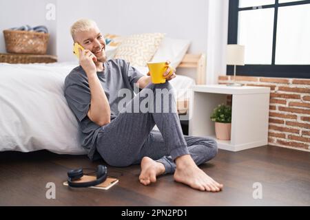 Junger weißer Mann, der mit dem Smartphone spricht und Kaffee im Schlafzimmer trinkt Stockfoto