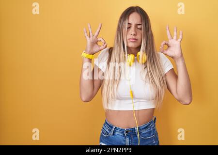 Junge blonde Frau, die über gelbem Hintergrund steht, Kopfhörer trägt, entspannt und mit geschlossenen Augen lächelt und mit den Fingern Meditationsgeste macht. Yoga Stockfoto