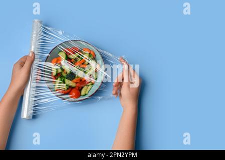 Weibliche Hände wickeln Schüssel mit Salat in Stretchfolie auf blauem Hintergrund Stockfoto