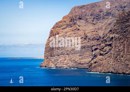 Majestätische Klippen von Acantilados de Los Gigantes stehen hoch, während ein kleines Segelboot vorbeifährt, mit der Landzunge Punta de Teno und dem Leuchtturm Teno. Stockfoto