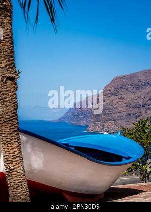 Klippen von Los Gigantes dominieren den Horizont auf diesem Foto, das vom Aussichtspunkt Mirador Archipenque mit einem alten Holzboot unter einer Palme aufgenommen wurde. Stockfoto