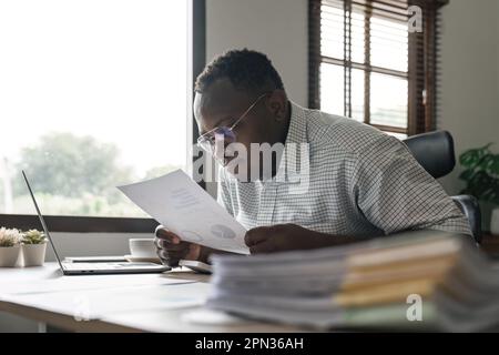 Fokussierter, seriöser afroamerikanischer Buchhalter, der Dokumente hält, während er einen Laptop-Bildschirm für Online-Handelstechnik verwendet Stockfoto