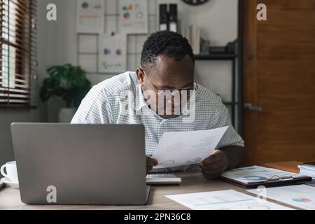 Fokussierter, seriöser afroamerikanischer Buchhalter, der Dokumente hält, während er einen Laptop-Bildschirm für Online-Handelstechnik verwendet Stockfoto