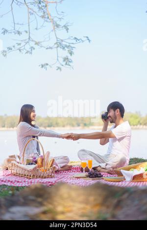 Ein verliebtes junges Paar, das sich die Hände zusammenhält und ein Foto macht, während es sich auf die Decke setzt, um ein Picknick zu machen. Stockfoto